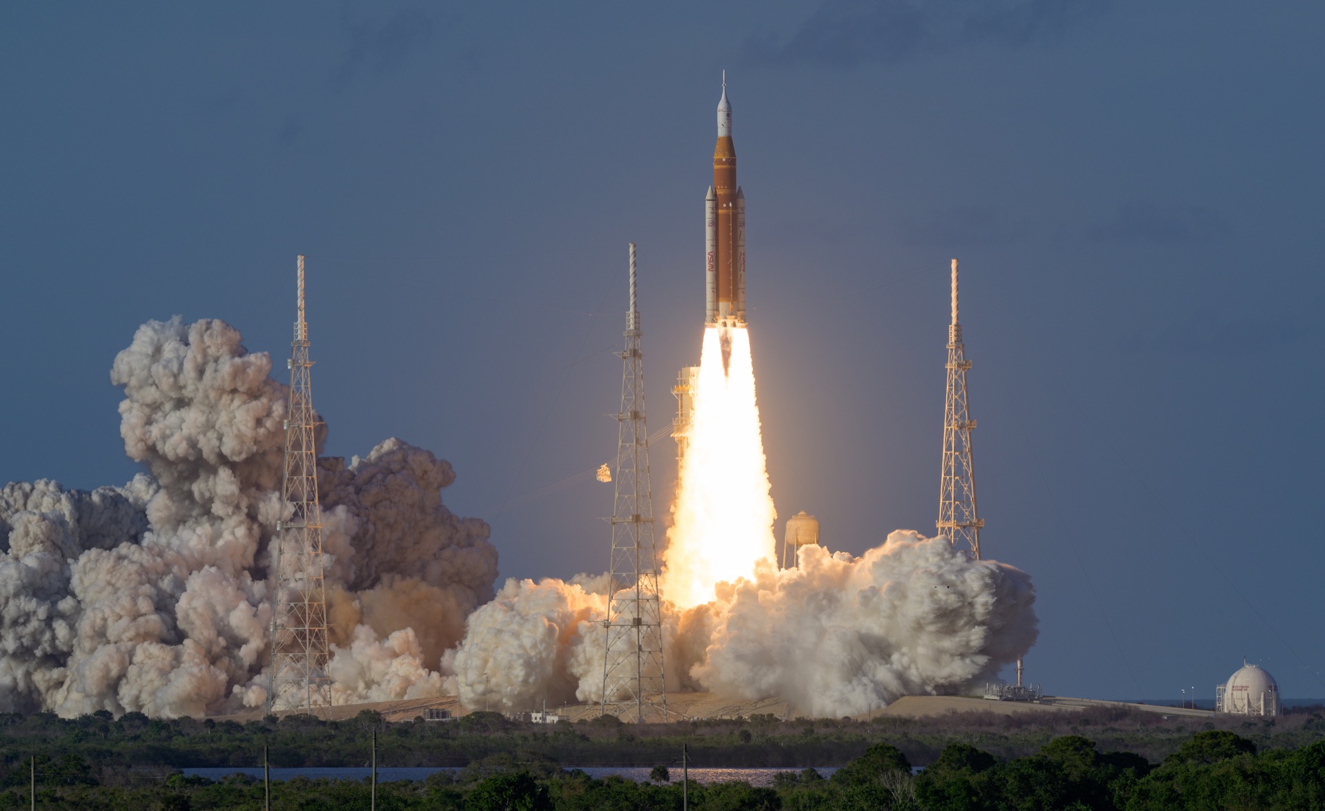 NASA's SLS rocket carrying the Orion spacecraft rises between two steel lightning towers at Kennedy Space Center, seconds after liftoff. The orange core stage glows warm against the deep blue-black night. Below the rocket, a blinding column of white-gold flame pours from the twin boosters, so bright it turns the launch pad into artificial daylight. Enormous clouds of steam — taller than the trees lining the shore — tumble outward in every direction, swallowing the base of the tower and rolling toward the dark Florida treeline in the foreground. The exhaust lights the clouds from within, giving them a warm amber glow at the edges that fades to grey. Above it all, the small white tip of Orion sits calmly atop the fury, pointed at the Moon.